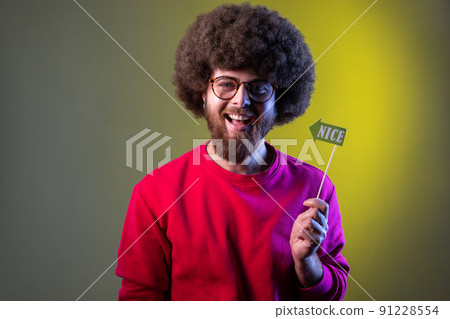 Happy positive hipster man with Afro hairstyle having event, holding party props with nice word in hands, wearing red sweatshirt. Indoor studio shot isolated on colorful neon light background. Happy positive hipster man with Afro hairstyle having event, holding party props with nice word in hands, wearing red sweatshirt. Indoor studio shot isolated on colorful neon light background. 91228554