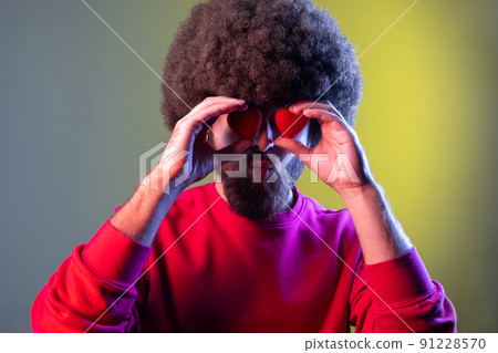 Portrait of hipster man with Afro hairstyle covering eyes with small red hearts, expressing romantic feelings, wearing red sweatshirt. Indoor studio shot isolated on colorful neon light background. 91228570