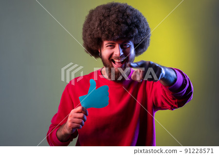Portrait of excited hipster man with Afro hairstyle holding and showing like or thumbs up paper shape sign, pointing finger to camera. Indoor studio shot isolated on colorful neon light background. 91228571