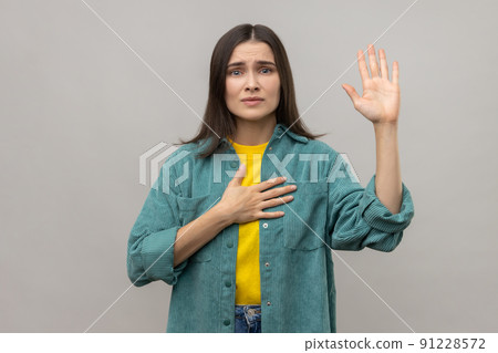 Serious patriotic woman holding hand on heart, juryman swearing to speak truth in court, honor and conscience, wearing casual style jacket. Indoor studio shot isolated on gray background. 91228572