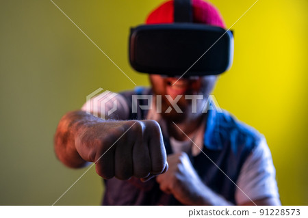 Hipster man in virtual reality glasses on head playing fighting game, holding clenched fists up to camera, ready to boxing. Indoor studio shot isolated on colorful neon light background. Hipster man in virtual reality glasses on head playing fighting game, holding clenched fists up to camera, ready to boxing. Indoor studio shot isolated on colorful neon light background. 91228573