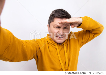 Portrait of serious middle aged man looking into distance with attentive view, point of view, keeps hand near forehead, wearing urban style hoodie. Indoor studio shot isolated on white background. 91228576