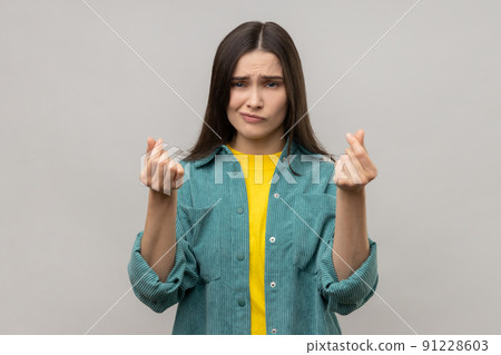 Portrait of beautiful woman makes money gesture, rubs fingers, looking at camera with glad expression, wearing casual style jacket. Indoor studio shot isolated on gray background. 91228603