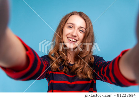 Beautiful woman wearing striped casual style sweater, making selfie POV, having positive expression, being in good mood, point of view. Indoor studio shot isolated on blue background. 91228684