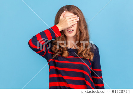 Don't want to look. Portrait of woman wearing striped casual style sweater standing covering eyes with hand, does not want see at something shameful. Indoor studio shot isolated on blue background. 91228734