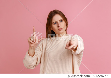 Portrait of serious blond woman pointing finger at you showing loser gesture, blaming and mocking, social pressure, wearing white sweater. Indoor studio shot isolated on pink background. 91228746