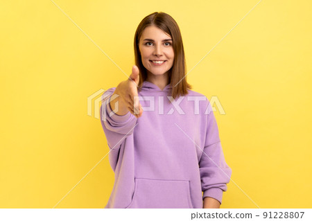 Nice to meet you. Portrait of pleased friendly smiling woman stretching her hand welcoming guests, hospitality, wearing purple hoodie. Indoor studio shot isolated on yellow background. Nice to meet you. Portrait of pleased friendly smiling woman stretching her hand welcoming guests, hospitality, wearing purple hoodie. Indoor studio shot isolated on yellow background. 91228807