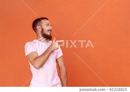 Portrait of optimistic bearded man standing, looking away with happy positive face and pointing at wall copyspace, wearing pink T-shirt. Indoor studio shot isolated on orange background. 91228823