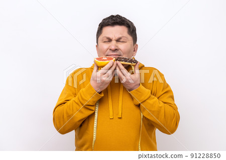 Man holding in hands half of juicy grapefruit and round donut with chocolate, enjoying fresh aroma of ripe fruit and sweets, wearing urban style hoodie. Indoor studio shot isolated on white background 91228850