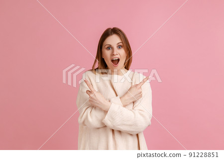 Portrait of beautiful amazed blond woman standing with crossed hands and pointing to copy space on both sides, wearing white sweater. Indoor studio shot isolated on pink background. 91228851