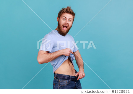 Portrait of bearded man having willpower points at slim waist in big trousers, successful weight loss, keeping diet and going for sport. Indoor studio shot isolated on blue background. 91228931