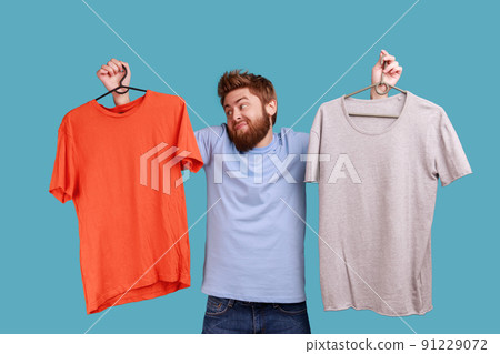 Portrait of puzzled doubtful handsome bearded man holding two hangers with gray and orange T-shirts, choosing outfit for going for a walk. Indoor studio shot isolated on blue background. Portrait of puzzled doubtful handsome bearded man holding two hangers with gray and orange T-shirts, choosing outfit for going for a walk. Indoor studio shot isolated on blue background. 91229072