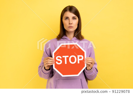 Portrait of serious attractive woman holding red stop sign, looking at camera strict expression, prohibition, wearing purple hoodie. Indoor studio shot isolated on yellow background. 91229078
