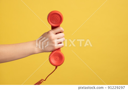 Profile side view closeup of woman hand holding and showing red call telephone handset receiver, call center. Indoor studio shot isolated on yellow background. Profile side view closeup of woman hand holding and showing red call telephone handset receiver, call center. Indoor studio shot isolated on yellow background. 91229297