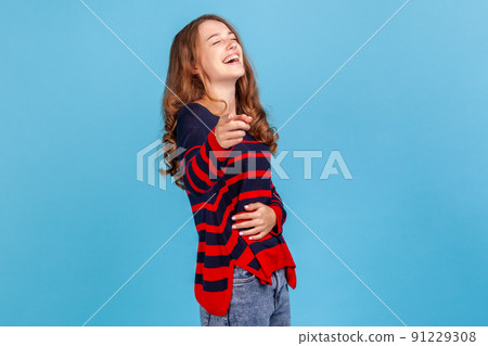 Lol, hey you. Excited joyful woman wearing striped sweater laughing out loud, holding her belly and pointing finger to camera, making fun of people. Indoor studio shot isolated on blue background. 91229308