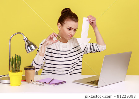 Dissatisfied woman sitting on workplace showing gold bitcoin and arrow pointing down to laptop display, downgrade of electronic currency. Indoor studio studio shot isolated on yellow background. Dissatisfied woman sitting on workplace showing gold bitcoin and arrow pointing down to laptop display, downgrade of electronic currency. Indoor studio studio shot isolated on yellow background. 91229398