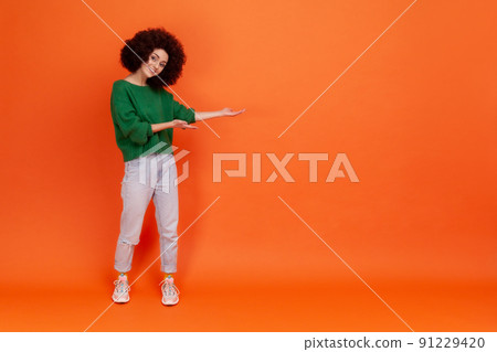 Full length portrait of smiling woman with Afro hairstyle wearing green casual style sweater presenting copy space for promotional text. Indoor studio shot isolated on orange background. 91229420