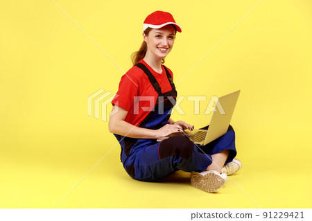 Side view portrait of satisfied handy woman sitting on floor with crossed legs and working on laptop, looking at camera, wearing overalls and red cap. Indoor studio shot isolated on yellow background. Side view portrait of satisfied handy woman sitting on floor with crossed legs and working on laptop, looking at camera, wearing overalls and red cap. Indoor studio shot isolated on yellow background. 91229421