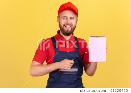 Optimistic smiling workman wearing blue overalls and red cap, holding pointing at paper notebook with pleasant smile, presenting copy space. Indoor studio shot isolated on yellow background. Optimistic smiling workman wearing blue overalls and red cap, holding pointing at paper notebook with pleasant smile, presenting copy space. Indoor studio shot isolated on yellow background. 91229430