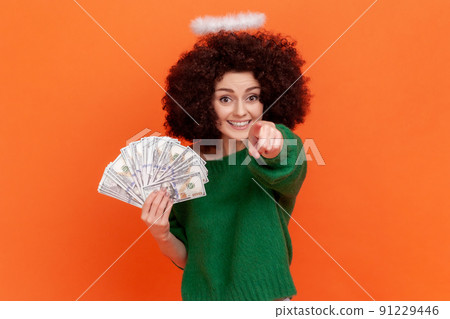 You are winner. Positive woman with Afro hairstyle in green casual sweater holding fan of dollars bills, pointing at you, choosing person who wins. Indoor studio shot isolated on orange background. You are winner. Positive woman with Afro hairstyle in green casual sweater holding fan of dollars bills, pointing at you, choosing person who wins. Indoor studio shot isolated on orange background. 91229446