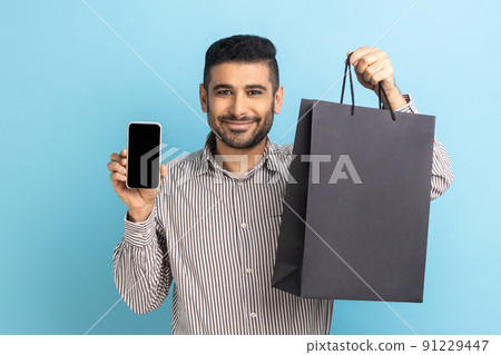 Smiling businessman with beard holding white display smartphone and black paper bag, online shopping, fast delivery, marketplace, wearing striped shirt. Indoor studio shot isolated on blue background. 91229447