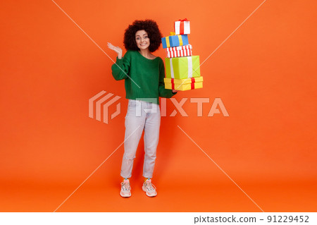 Full length portrait of happy woman with Afro hairstyle wearing green casual style sweater holding many present boxes, spreading palm aside. Indoor studio shot isolated on orange background. Full length portrait of happy woman with Afro hairstyle wearing green casual style sweater holding many present boxes, spreading palm aside. Indoor studio shot isolated on orange background. 91229452
