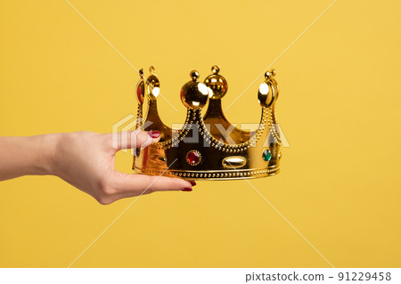 Closeup of woman hand holding golden crown, concept of awards ceremony, privileged status, superior position. Indoor studio shot isolated on yellow background. 91229458