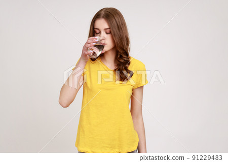 Portrait of attractive thirsty woman of young age with brown wavy hair in yellow casual T-shirt drinking water from glass, water balance. Indoor studio shot isolated on gray background. Portrait of attractive thirsty woman of young age with brown wavy hair in yellow casual T-shirt drinking water from glass, water balance. Indoor studio shot isolated on gray background. 91229483