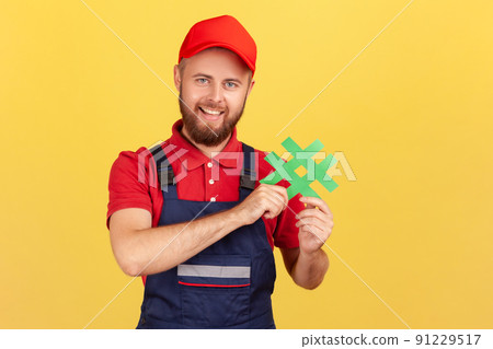 Portrait of positive delighted workman wearing blue overalls standing, holding green hashtag, looking at camera with happy expression. Indoor studio shot isolated on yellow background. Portrait of positive delighted workman wearing blue overalls standing, holding green hashtag, looking at camera with happy expression. Indoor studio shot isolated on yellow background. 91229517