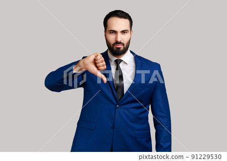 Bearded man criticizing bad quality with thumbs down displeased grimace, showing dislike gesture, expressing disapproval , wearing official style suit. Indoor studio shot isolated on gray background. Bearded man criticizing bad quality with thumbs down displeased grimace, showing dislike gesture, expressing disapproval , wearing official style suit. Indoor studio shot isolated on gray background. 91229530
