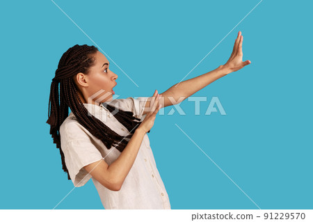 Side view portrait of frightened woman tries to protect herself from something terrible, notices awful thing, makes stop gesture, stares with big eyes. Indoor studio shot isolated on blue background. Side view portrait of frightened woman tries to protect herself from something terrible, notices awful thing, makes stop gesture, stares with big eyes. Indoor studio shot isolated on blue background. 91229570