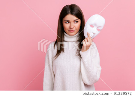 Portrait of young beautiful adult female with dark hair looking away, holding in hands white mask, wearing white casual style sweater. Indoor studio shot isolated on pink background. Portrait of young beautiful adult female with dark hair looking away, holding in hands white mask, wearing white casual style sweater. Indoor studio shot isolated on pink background. 91229572
