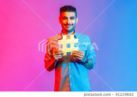 Portrait of man in shirt with toothy smile on his face holding hashtag symbol, recommending popular topics, internet trends. Indoor studio shot isolated on colorful neon light background. 91229682