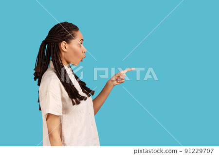 Side view of surprised woman with black dreadlocks pointing away with finger, showing blank space for your advertisement, wearing white shirt. Indoor studio shot isolated on blue background. Side view of surprised woman with black dreadlocks pointing away with finger, showing blank space for your advertisement, wearing white shirt. Indoor studio shot isolated on blue background. 91229707