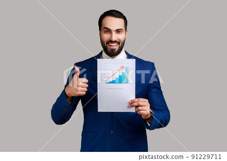 Handsome bearded businessman standing showing business growth graph, looking at camera, showing thumb up, wearing official style suit. Indoor studio shot isolated on gray background. Handsome bearded businessman standing showing business growth graph, looking at camera, showing thumb up, wearing official style suit. Indoor studio shot isolated on gray background. 91229711