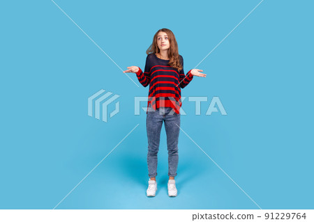Full length portrait of uncertain woman wearing striped sweater looking desperate, making helpless gesture with her hands, doesn't know what to do. Indoor studio shot isolated on blue background. 91229764