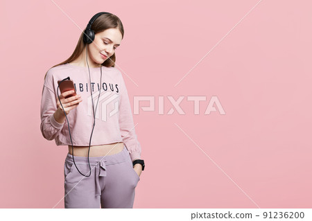 Adorable slim young woman in casual outfit enjoys favourite audio track in hearphones, uses modern cell phone, keeps hand in pocket, isolated over pink background with copy space for your text 91236200