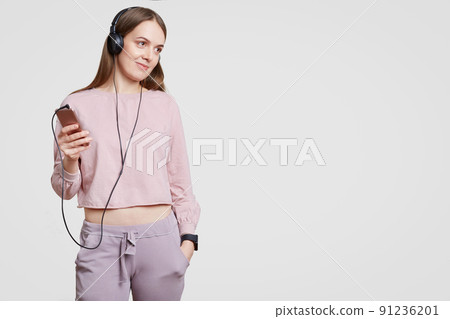 Studio shot of pleased attractive woman dressed in casual clothes, keeps hand in pcket, holds modern electronic gadget, listens favourite music in headphones, isolated over white background. 91236201