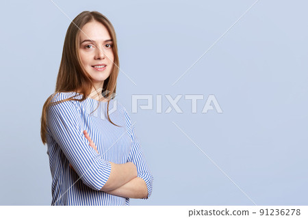 Sideways portrait of glad beautiful young woman with long dark hair wears ellegant striped blouse, keeps hands crossed, poses against blue background with copy space for your promotional text 91236278
