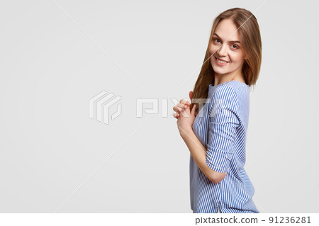 Studio shot of pleased brunette woman with appealing look, dressed in stylish outfit, smiles positively at camera, poses for fashion magazine, isolated over white background with free space. 91236281