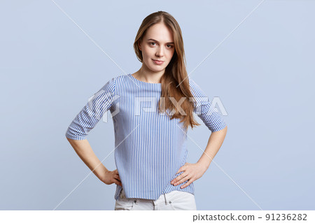 Horizontal shot of pretty female in stylish clothing keeps hands on waist, looks with confident and determined expressionn in camera, poses against blue background. Woman being sure in herself 91236282