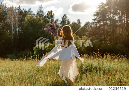 Young beautiful woman, wearing white dress, holding flowers and dancing on the meadow. Girl joying nature and freedom. Natural beauty. Dance, movement. Mental health, stress free, dreaming. Sunset. Young beautiful woman, wearing white dress, holding flowers and dancing on the meadow. Girl joying nature and freedom. Natural beauty. Dance, movement. Mental health, stress free, dreaming. Sunset. 91236519
