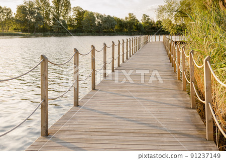 Long Wooden bridge over lake. Beautiful sunrise over lake pier. Planked footway on the river or lake Long Wooden bridge over lake. Beautiful sunrise over lake pier. Planked footway on the river or lake 91237149