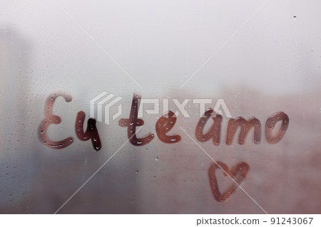 Lettering Portuguese text Eu te amo I love you in english and heart shape on sunset wet window 91243067
