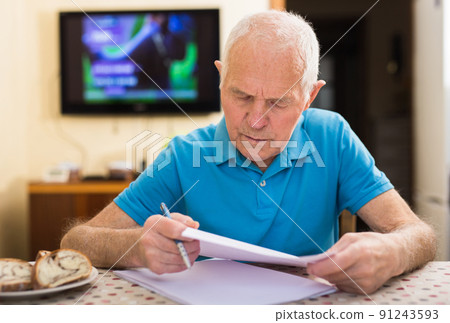 Elderly man writes a letter on a sheet of paper while sitting at a table in room Elderly man writes a letter on a sheet of paper while sitting at a table in room 91243593