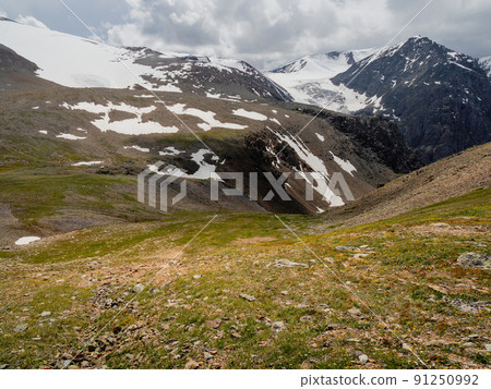 Summer view from sunlitgreen rocky slope to a narrow mountain gorge under cloudy sky. Colorful landscape with large snow mountains at changeable weather. 91250992