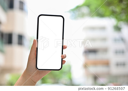 A smartphone white screen mockup in a female's hand over blurred flat or apartment building 91268507