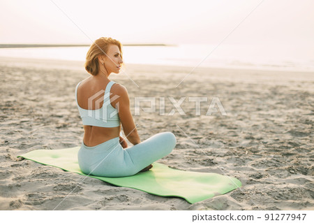 A beautiful girl is sitting on the seashore in the lotus position and resting after yoga or fitness classes. Healthy lifestyle, self-care, activity 91277947