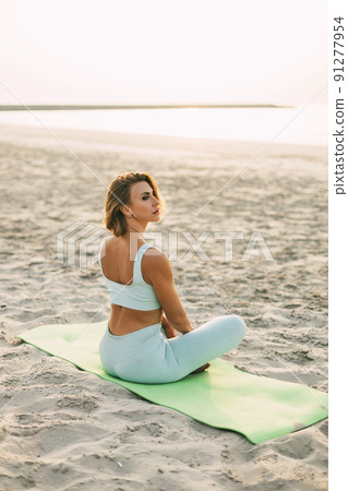 A beautiful girl is sitting on the seashore in the lotus position and resting after yoga or fitness classes. Healthy lifestyle, self-care, activity 91277954