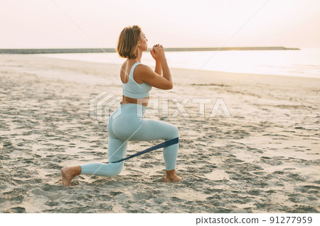 A sporty woman trains with a resistance band, makes lunges.A girl in sportswear trains on the beach A sporty woman trains with a resistance band, makes lunges.A girl in sportswear trains on the beach 91277959